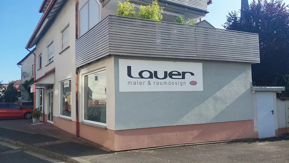 Ein zweistöckiges Gebäude mit einem großen Schild an der Seite, auf dem „Lauer Maler & Raumdesign“ steht. Über dem Schild befindet sich ein Balkon, die Fenster auf beiden Etagen sind geschlossen, und daneben parkt ein rotes Auto. - Firmenadresse